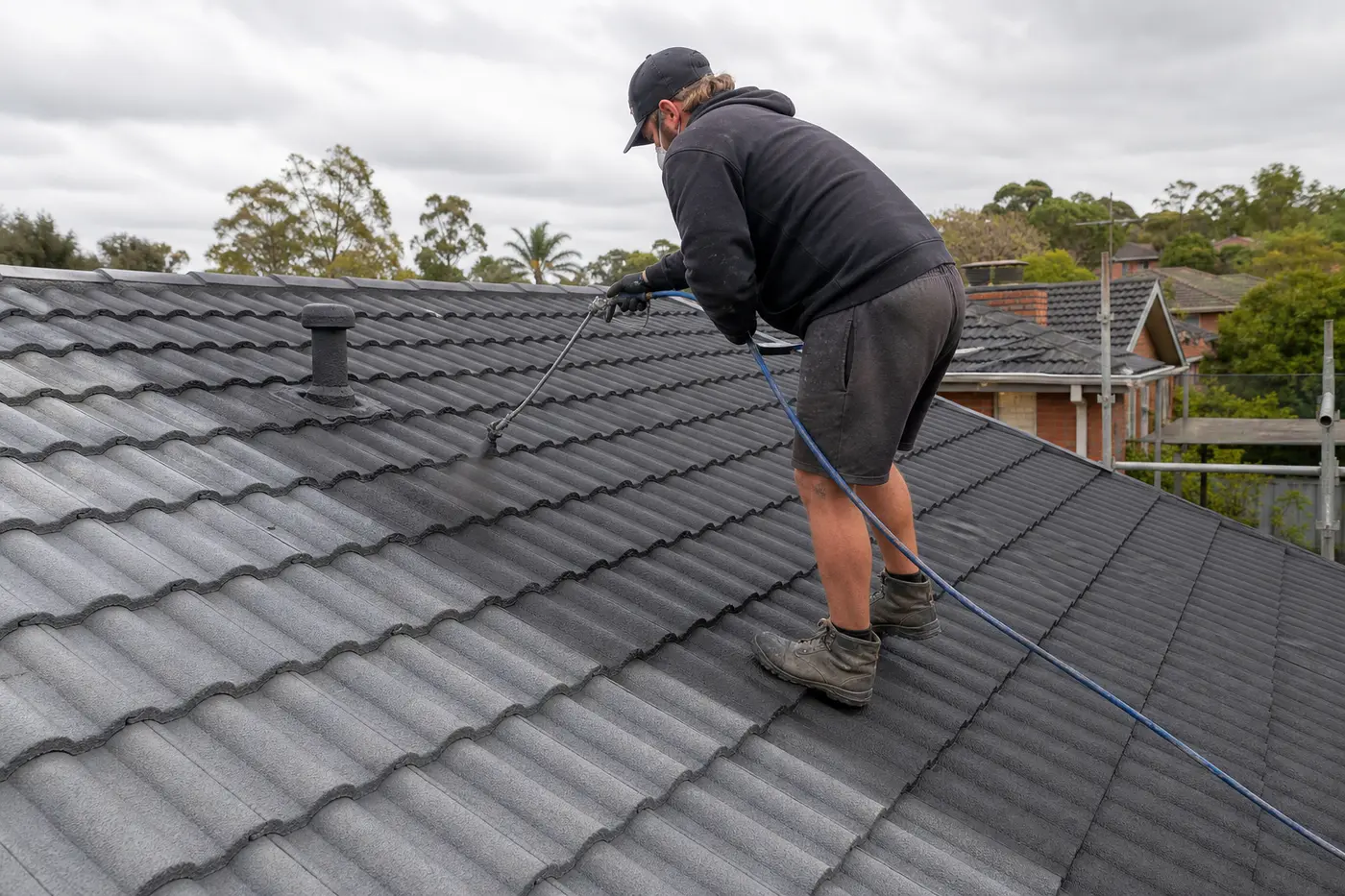 Roofer spray-painting a freshly cleaned concrete tile roof deep charcoal on a single-storey northern-suburbs Melbourne home, seen from roof level